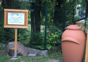 Zbiornik na deszczówkę i tablica informacyjna z tekstem: Utworzenie punktu dydaktycznego zrealizowane przy udziale środków Wojewódzkiego Funduszu Ochrony Środowiska i Gospodarki Wodnej w Łodzi w ramach konkursu "Nasze ekologiczne pracownie"