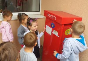 Chłopiec wrzuca list do dużej, czerwonej skrzynki. Pięcioro dzieci się przygląda.