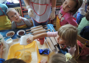 Dzieci zgromadzone wokół stolika z przyborami do wykrawania ciastek. Dziewczynka wsypuje cukier do miski.