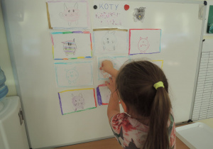 Druga dziewczynka przywiesza rysunek kota na tablicy.
