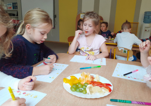 Dzieci przy stoliku próbują owoce i określają ich smak w kartach pracy.