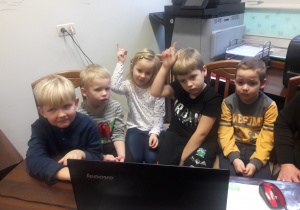 Pięcioro dzieci przed komputerem. Chłopiec i dziewczynka podnoszą ręce.