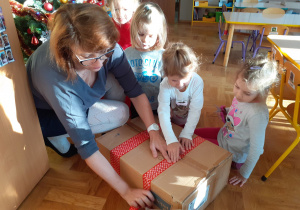 Nauczycielka w towarzystwie dzieci okleja karton czerwoną taśmą.
