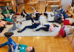 Dzieci leżą na dywanie na plecach i podnoszą do góry jedną nogę.