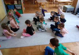 Dzieci siedzą skrzyżnie na dywanie. Wykonują ćwiczenia rozciągające z rękami za plecami.