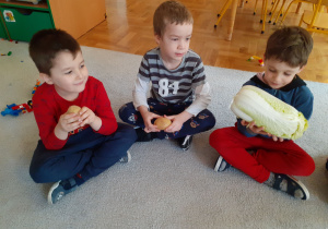 Trzech chłopców siedzi na dywanie trzymając w rękach warzywa.