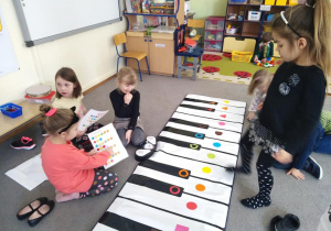 Dziewczynka gra na macie muzycznej, a druga wskazuje kolejne elementy w instrukcji.