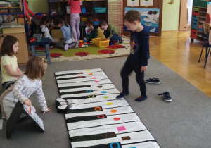 Chłopiec gra na macie muzycznej według instrukcji pokazywanej przez koleżankę.