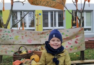 Chłopiec siedzi nadrewnianej palecie. Obok niego znajdują się jesienne warzywa i owoce. Za nim na drzewach wiszą drewniane łyżki, tło w kolorowe plamy na papierze pakowym i napis "fotobudka 2021/2022".