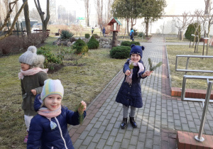 Dzieci chodzą po ogrodzie. Dwie dziewczynki pokazują gałęzie świerku.