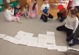 Dzieci siedzą na dywanie. Przed nimi kartki z zapisanymi liczbami i działaniami ułożonymi pod liczbą wskazującą ich wynik.