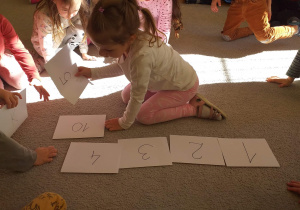 Dzieci układają na dywanie kartki z zapisanymi liczbami od 1 do 10.