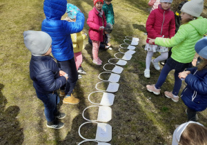 Na trawie leżą papierowe koszyczki z zapisanymi liczbami i kostką z ilością oczek obrazującą tę liczbę. Po jednej i drugiej stronie stoją dzieci.
