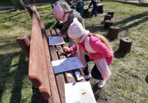 Na ławce leżą kartki, kredki ołówki. Przed nimi kucają dzieci. Przygotowują wielkanocne prace plastyczne wykorzystując zebrane w ogrodzie pisanki.
