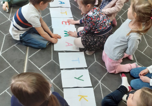 Dzieci zgromadzone na dywanie wokół napisu "matematyka" ułożonego z liter zapisanych na kartkach formatu A4.