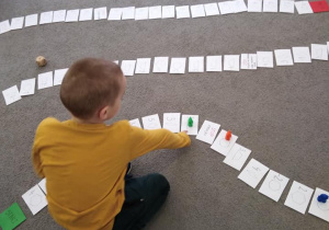 Na dywanie leżą ułożone kolejno kartoniki z zadaniami i figurami geometrycznymi. Chłopiec wskazuje kartonik, na którym stoi zielony pionek.