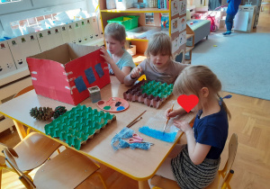 Trzy dziewczynki przy stoliku malują kartonowe pudełko, wytłoczkę na jajka i folię bąbelkową.