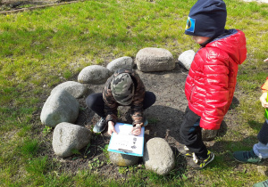Chłopiec kuca przy kamieniach i zapisuje coś na kartce przyczepionej do podkładki.