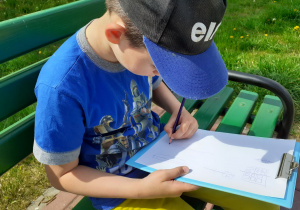Chłopiec siedzi na ławce i rysuje na kartce umieszczonej na podkładce.