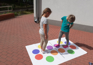 Dwie dziewczynki na tarasie stoją na planszy z kolorowymi kółkami.