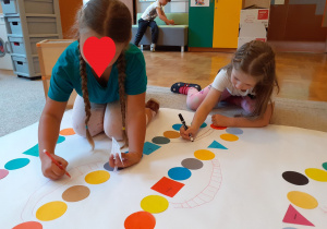 Dziewczynki rysują drabiny między kolorowymi figurami geometrycznymi na planszy.