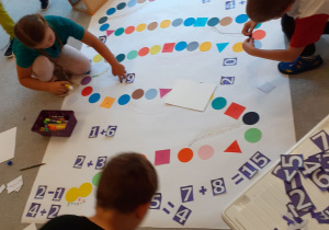 Dzieci przyklejają liczby i działania na planszy do konstruowanej gry planszowej.