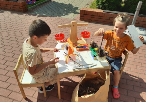 Dwaj chłopcy ważą szyszki i patyki, siedząc przy stoliku na tarasie.