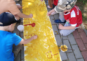 Dzieci malują patyczkami umoczonymi w wodzie z proszkiem do prania na długim kartonie pomalowanym kurkumą.