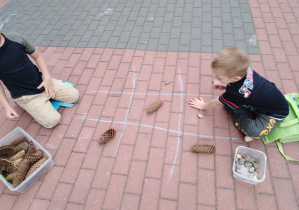 Dwaj chłopcy grają w "kółko i krzyżyk", układając szyszki i kamienie na planszy narysowanej kredą na tarasie.