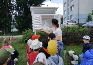 Dzieci wraz z nauczycielką zaglądają do stacji meteorologicznej w celu odczytania pomiaru temeratury i ciśnienia atmosferycznego.
