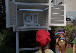 Dzieci zaglądają do stacji meteorologicznej, by odczytać wartość temeratury i ciśnienia atmosferycznego.