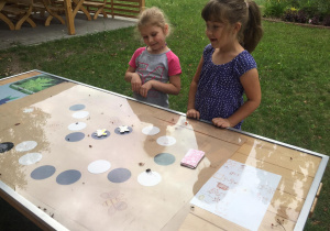 Dwie dziewczynki przy ławostole, na którym umieszczona została zrobiona przez dzieci gra planszowa.