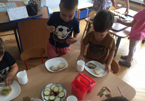 Chłopcy siadają do stołu, na którym stoją talerze z posmarowanym chlebem, kubki i talerze z produktami na kanapki. Na jednym z talerzy są pomidorki, pokrojona cukinia i jeżyny zebrane przez dzieci w ogrodzie.