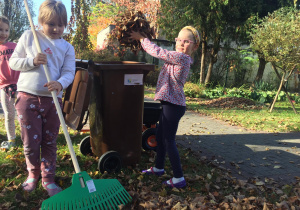 Dziewczynka grabi liście, a druga wrzuca liście do pojemnika.