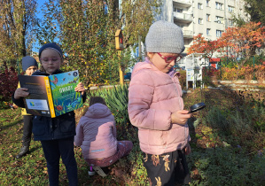 Dzieci poszukują w ogrodzie owadów korzystając z lupy i książek o tematyce przyrodniczej.