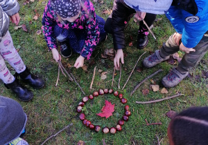 Dzieci układają mandalę z kasztanów i patyków wokół liścia.