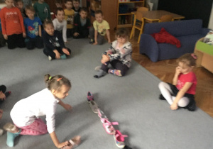 Dzieci siedzą na dywanie. Przed nimi ustawiony rząd butów. Z lewej strony dziewczynka kuca z butem w ręku.