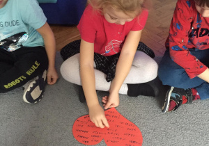 Dziewczynka siedząca skrzyżnie na dywanie, wbija szpilkę w czerwone papierowe serce z wypisanymi imionami.