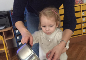 Dziewczynka z pomocą nauczycielki przelewa wosk z rondelka do miski przez klucz z folii aluminiowej.