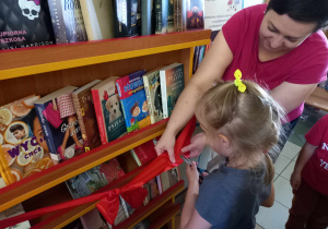Dziewczynka przecina czerwoną wstążkę przy regale z książkami. Pani Dyrektor podtrzymuje wstążkę.