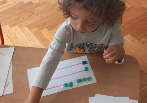 Chłopiec układa koraliki na podkładce z narysowanymi liniami poziomymi.