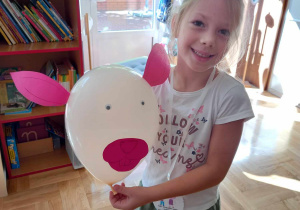 Dziewczynka trzyma w rękach biały balon z przyklejonymi różowymi uszami i pyskiem.