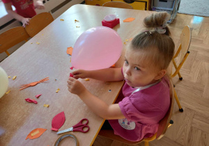 Dziewczynka siedzi przy stoliku. Obok niej leży różowy balon i papierowe uszy, nosy i wąsy do wycinania.