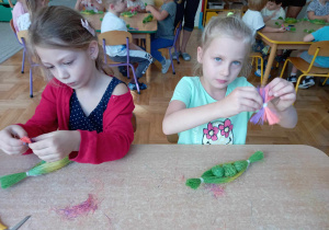 Dwie dziewczynki siedzą przy stoliku i formują kokardki z sizalowego sznurka.
