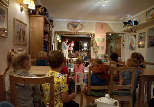 Dzieci siedzą na krzesełkach i oglądają przedstawienie, w którym aktorka trzyma kukiełkę dziewczynki.
