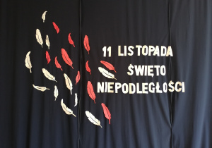 Dekoracja sali przedstawoająca napis 11 listopada Święto Niepodległości, białe i czerwone papierowe pióra na granatowym tle.
