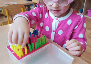 Dziewczynka w okularach wkłada kolorowe druciki w otwory w pudełku.