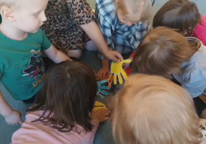 Dzieci siedzą w grupie na dywanie i pochylają się nad kolorowymi odciskami dłoni.