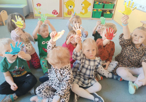 Dzieci siedzą na dywanie. Podnoszą do góry ręce, w których trzymają wycięte i zalaminowane kolorowe odciski dłoni.