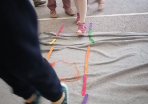 Dzieci przechodzą pomiędzy dwoma rzędami kolorowych patyków, omijając ułożoną pośrodku włóczkę.